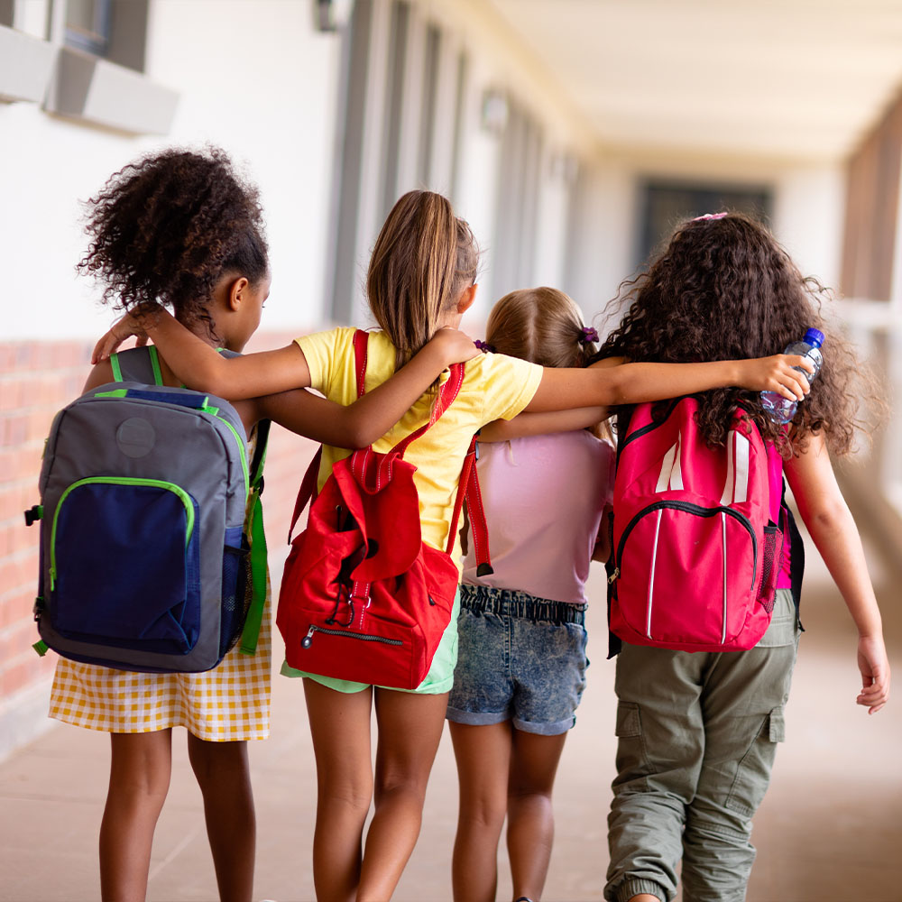 Four young girls with backpacks walking shoulder to shoulder with arms embracing each other