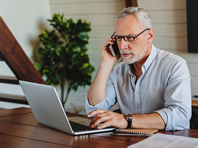 Older man with a serious expression talking on his smartphone and looking at his laptop