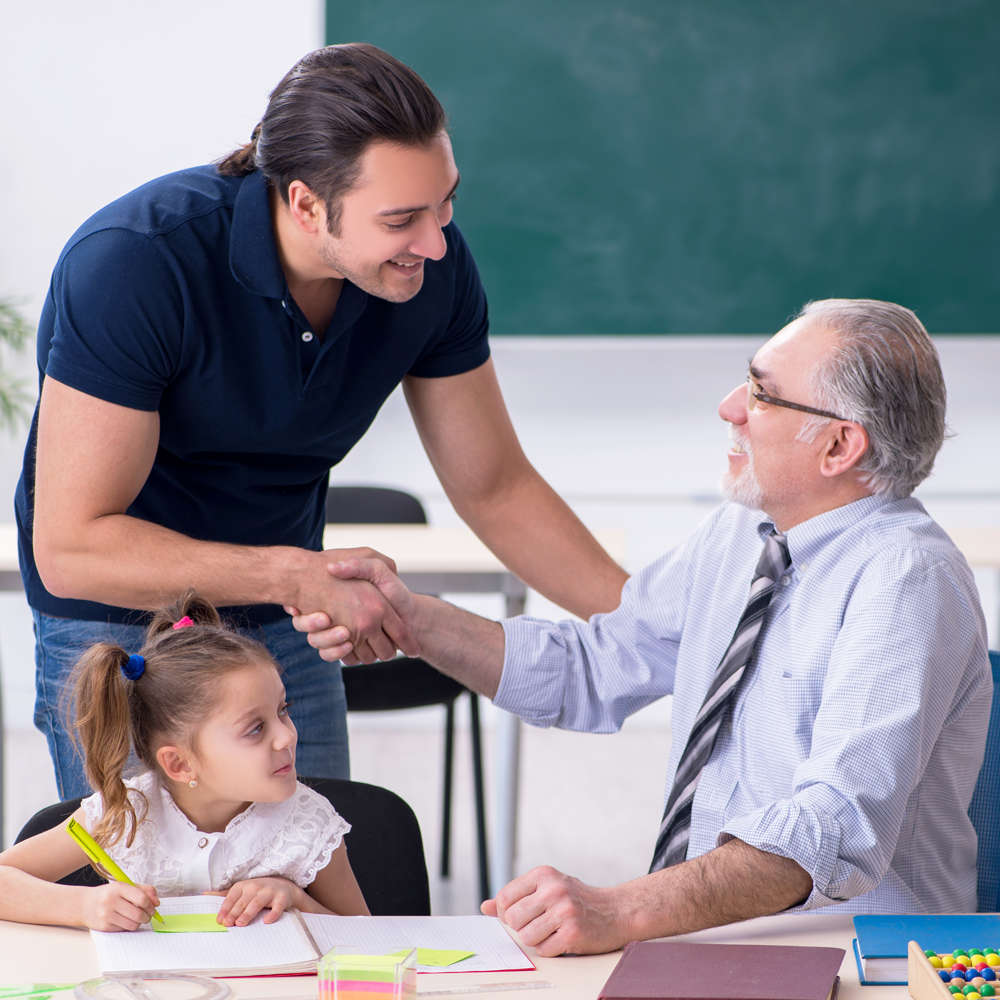 Parent shaking teacher's hand while child sits at desk writing in notebook