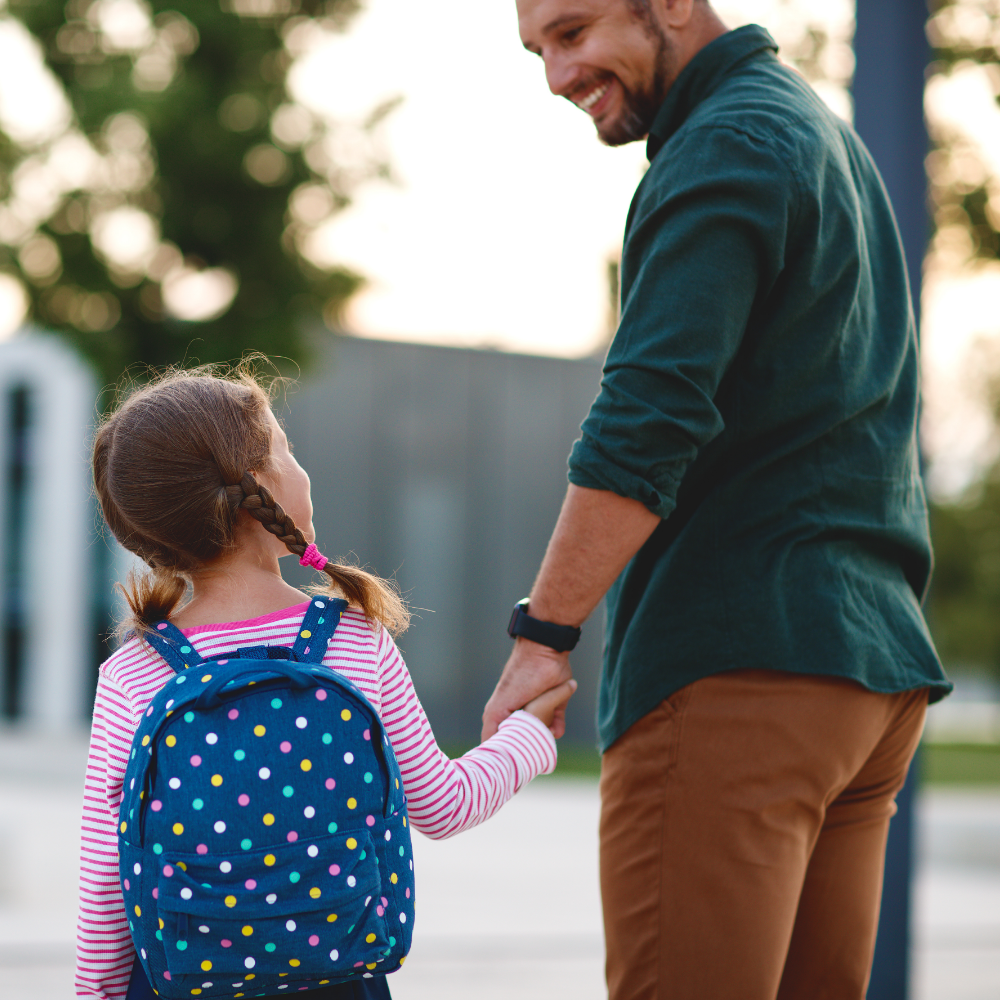 A father holding his backpack-wearing daughter's hand smiling at one another