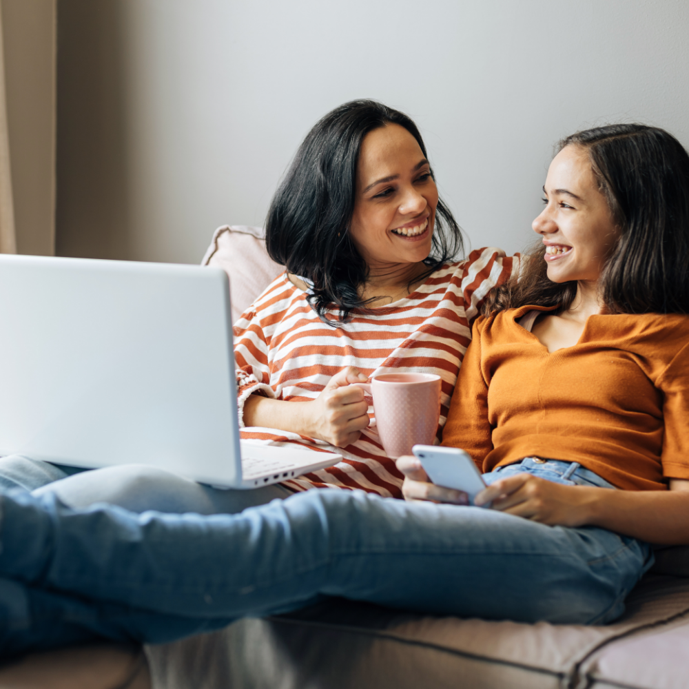 Mother and daughter sitting on a couch and smiling at one other while mother has a laptop and mug and daughter has her phone