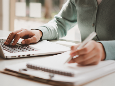Close-up of a woman typing on her computer and writing in a notepad