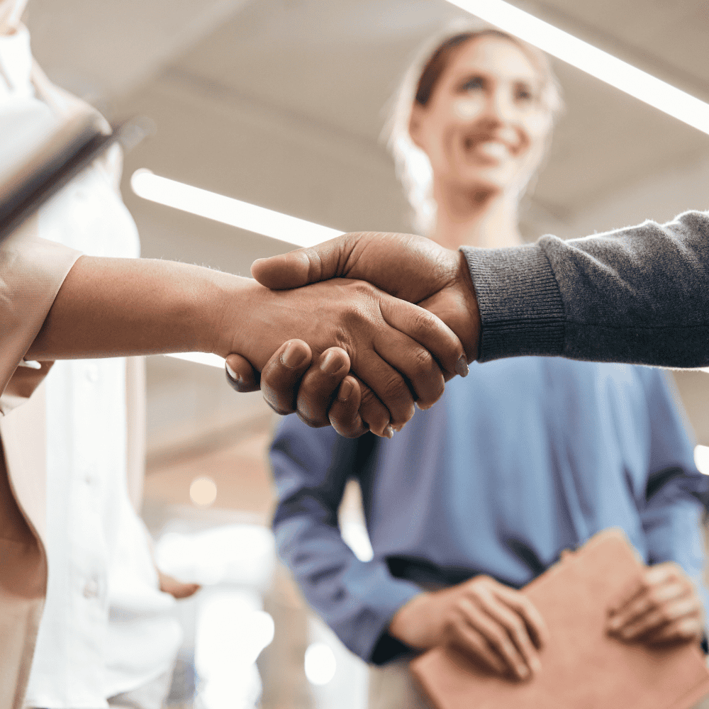 Close-up of a handshake between work colleagues with an out-of-focus woman smiling behind them in the workplace