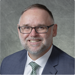 A bearded man, wearing glasses and smiling looks directly at the camera. He is professionally dressed in a suit and tie.