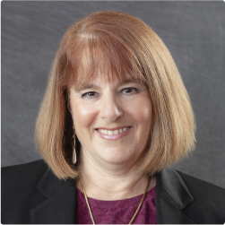 A professionally dressed woman with light colored, straight shoulder length hair is smiling and looking directly at the camera.